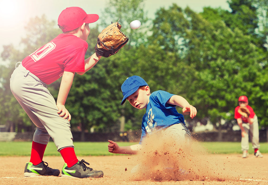 little league baseball team playing a game while the player in red is about to catch the ball while a player in blue slides in to the plate under him