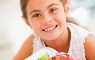 little girl smiling while holding a lunchbox of healthy food