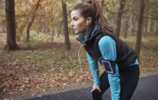 woman standing bent over with her hands on her knees while exercising in forest with workout clothes on and phone strapped to her arm