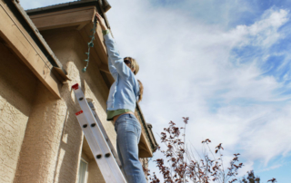 woman on a ladder stretching to hang lights on her home