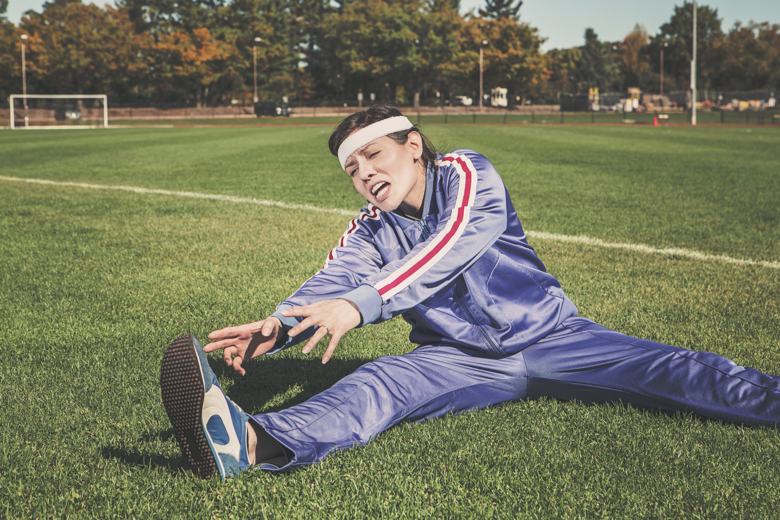 female wearing a blue track suit sitting on green sports field stretching with a pained look on her face