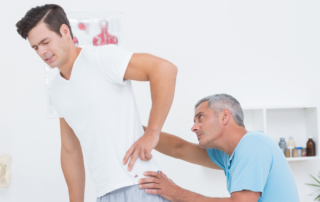 young male patient with an expression of pain on his face as he holds his hip is being looked at by an older male who is his physical therapist