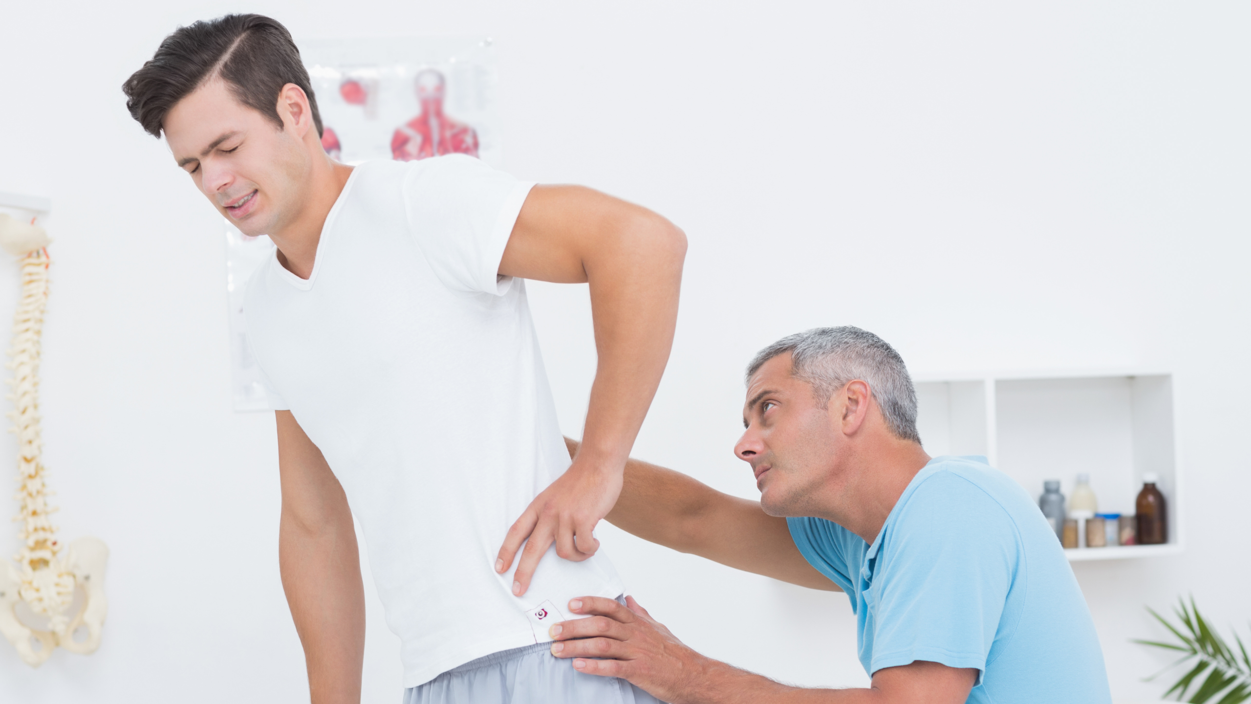 young male patient with an expression of pain on his face as he holds his hip is being looked at by an older male who is his physical therapist