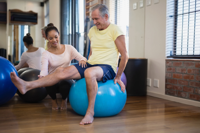 female physical therapist helping senior male patient doing leg exercise on blue ball at physical therapy appointment in san antonio