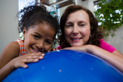 portrait of smiling girl and physical therapist leaning on fitness ball in clinic