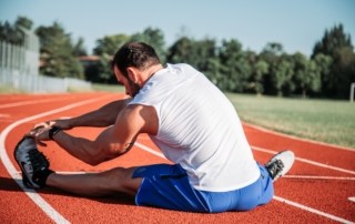man wearing athletic clothes and shoes sitting on a running track stretching to the left leg about to work out